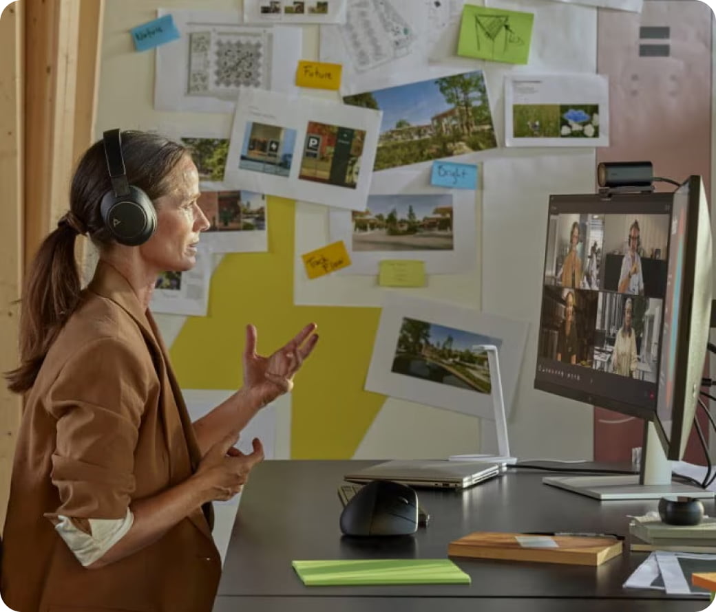 Une femme portant un micro-casque, assise à un bureau, faisant des gestes pendant un appel vidéo avec des collègues visibles à l’écran de son ordinateur. Derrière elle, un mur recouvert de photos, de notes et de croquis épinglés.