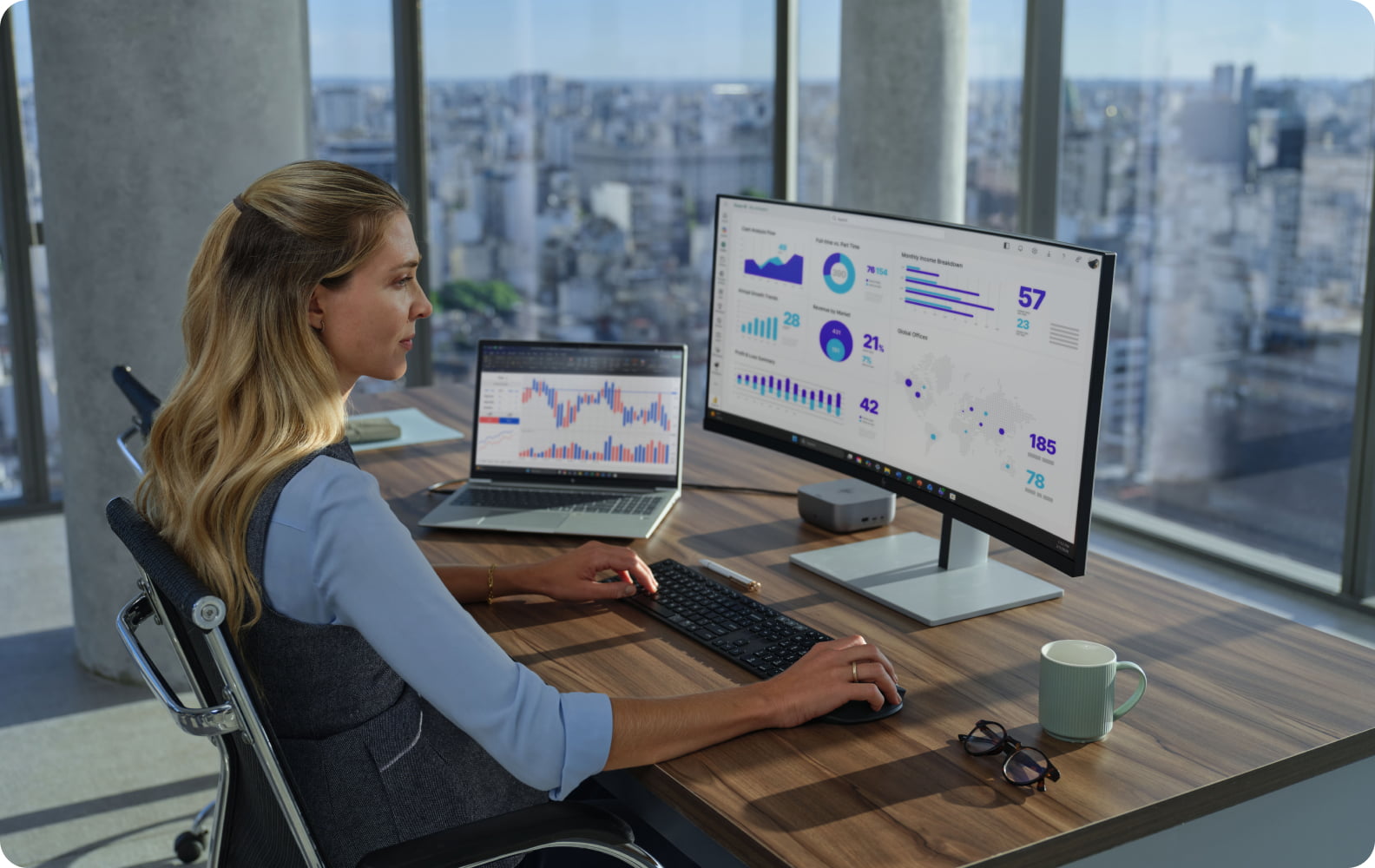 Une femme assise à un bureau dans un environnement de travail moderne, travaillant sur un PC fixe qui affiche des graphiques et des tableaux de données, un PC portable ouvert à côté d’elle. La ligne d’horizon de la ville est visible à travers de grandes fenêtres.