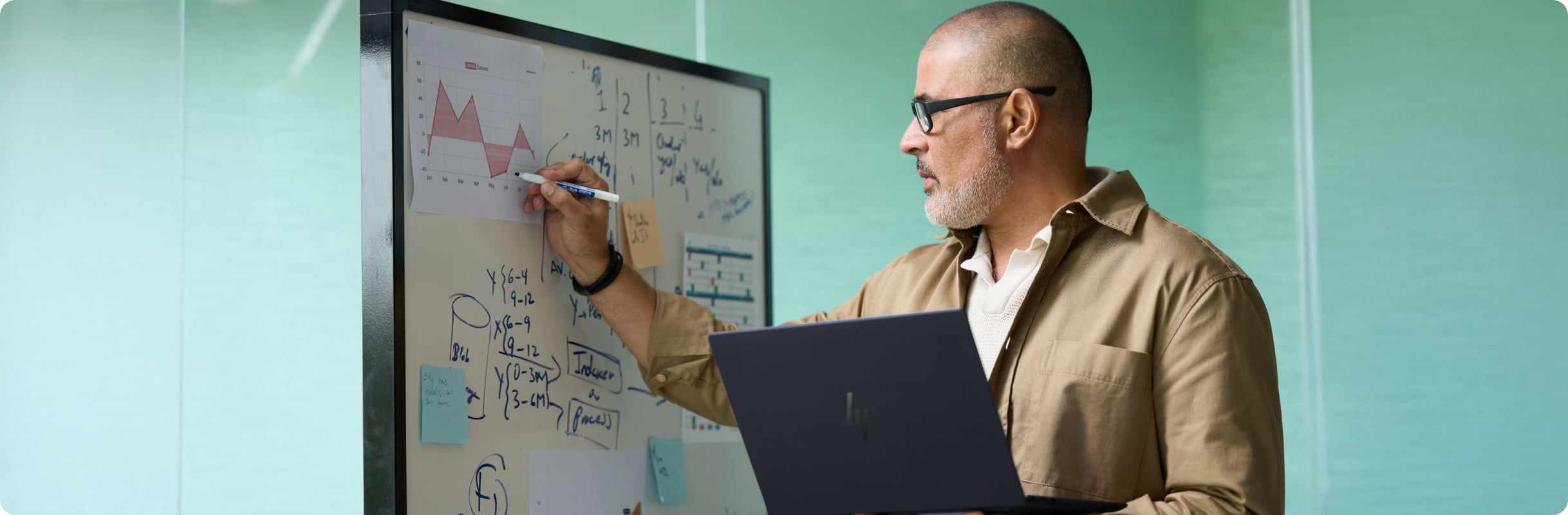 Professional standing at a whiteboard reviewing charts and handwritten notes while holding an HP laptop.
