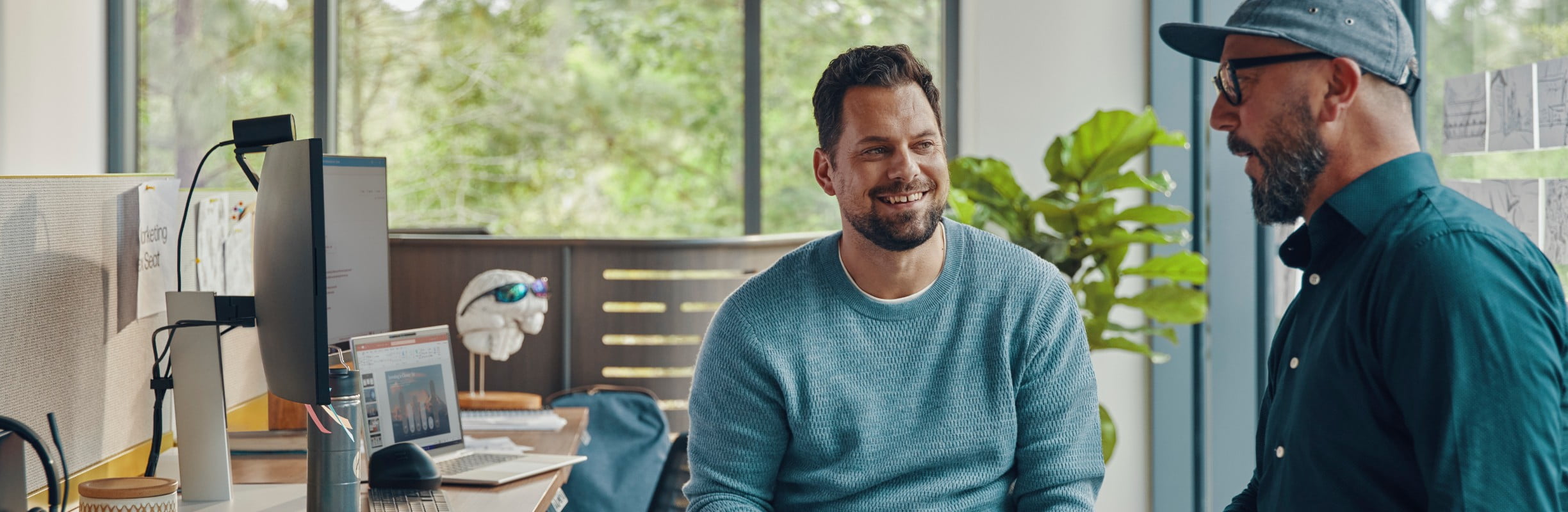 Dwaj mężczyźni rozmawiają przy biurku, na którym stoi laptop do firmy oraz monitor.