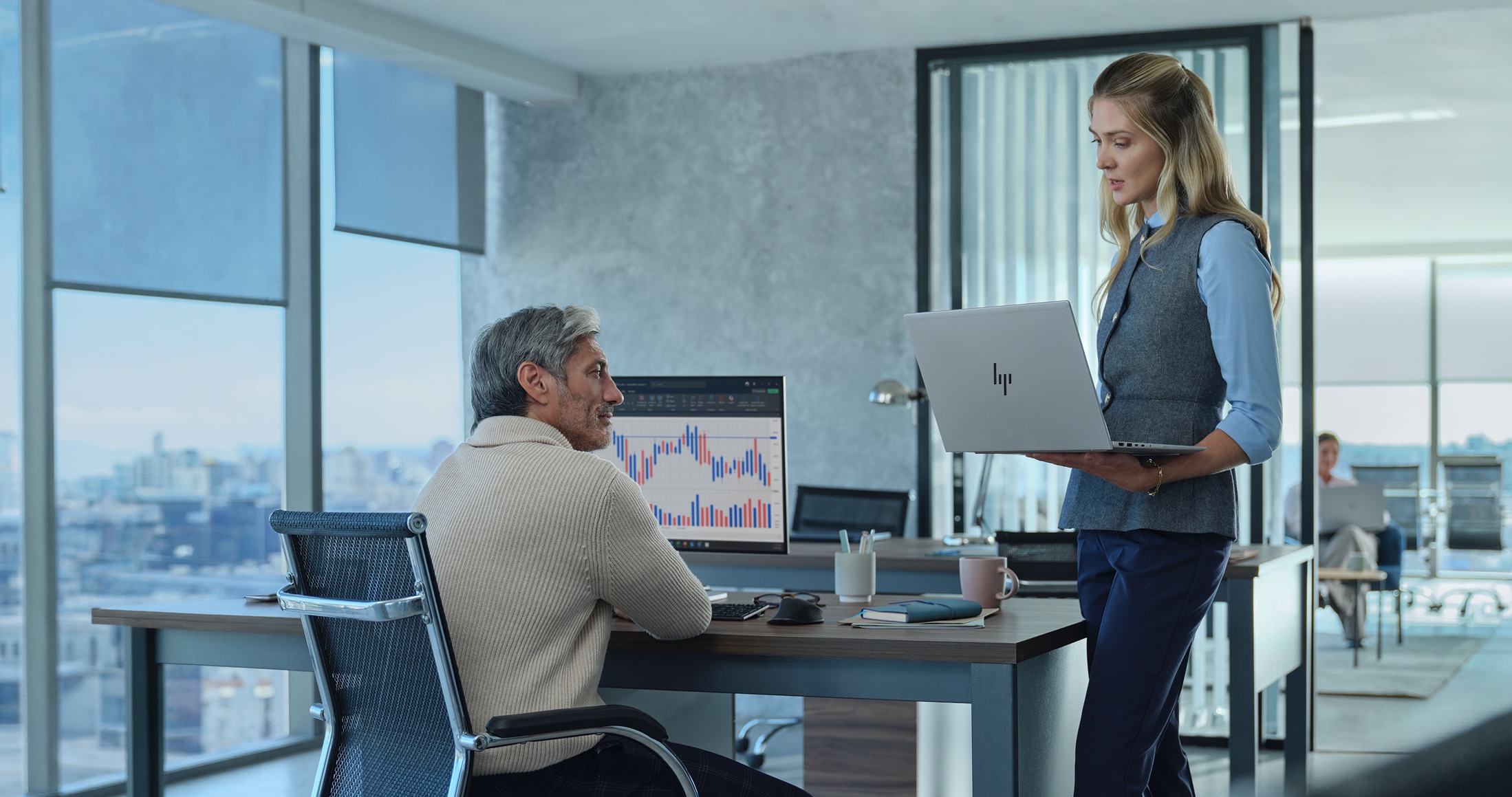 Man seated at a desk with a graph displayed on a monitor and looking up at a woman holding a laptop.