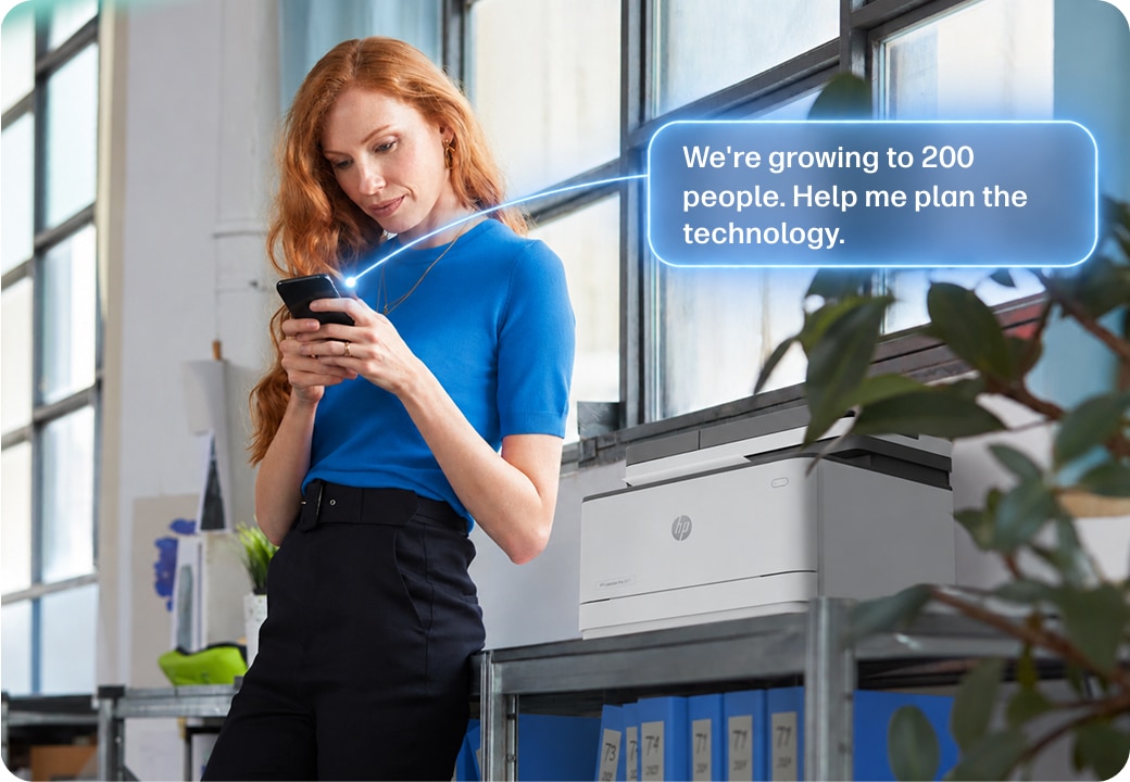 Person standing by a printer and typing on a smart phone with "We're growing to 200 people. Help me plan the technology." prompt in a blue bubble.
