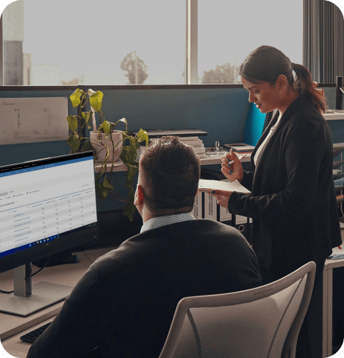A man at a desk reviews a spreadsheet on an HP monitor while a colleague stands beside him taking notes.