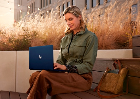Une femme travaillant dans son ordinateur portable HP sur ses genoux, assise sur un banc en bois.