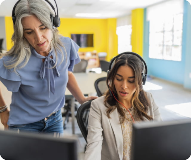 A woman standing beside a seated woman, both wearing headsets.