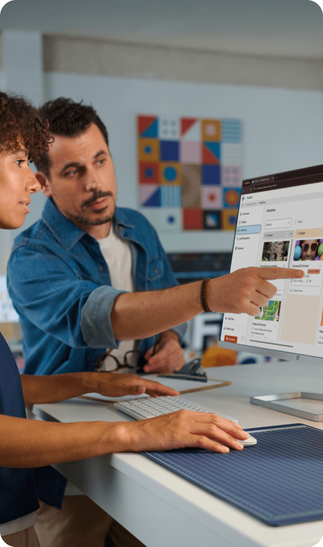 Two people collaborating at a desk while reviewing content on a large computer monitor.