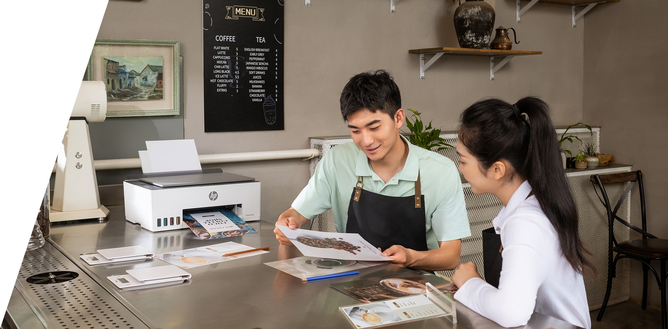 A wall with a shelf and a cabinet. An HP printer, toner cartridges, and printing paper are displayed on the cabinet, next to a photo frame. On the shelf are flowers, candlesticks, lamps, and photo frames.