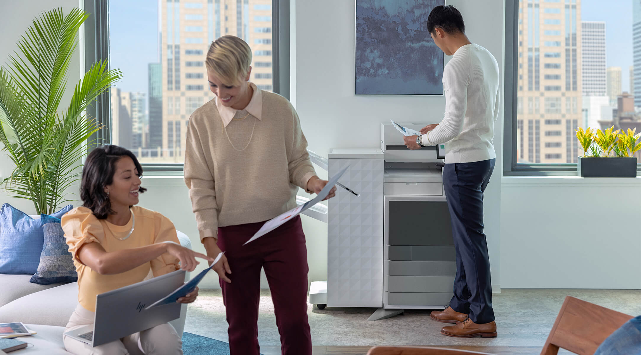 在一个现代化的办公室里，一位坐在沙发上的女士正在使用笔记本电脑，另一位站着的人正手持文件与她交流，背景中还有一位男士站在惠普智能复合机旁正在操作
