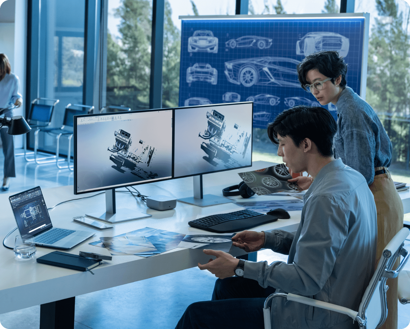 Man and woman in modern engineering office looking at car designs on an HP dual display and laptop with printed pages on the desk
