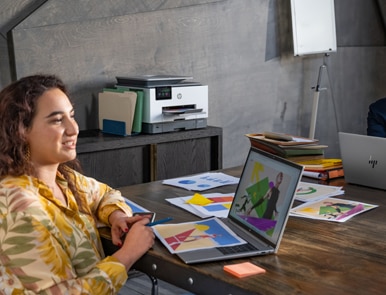 A woman sitting at a desk with colorful printed documents and an HP laptop, in a modern office setting with an HP printer in the background.
