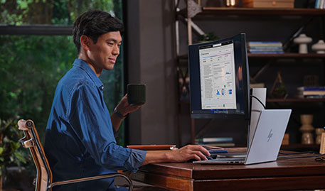 Man at a desk using a curved monitor and HP laptop, holding a coffee mug in a home office.