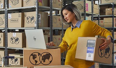 Woman in yellow sweater uses laptop among warehouse shelves filled with boxes.
