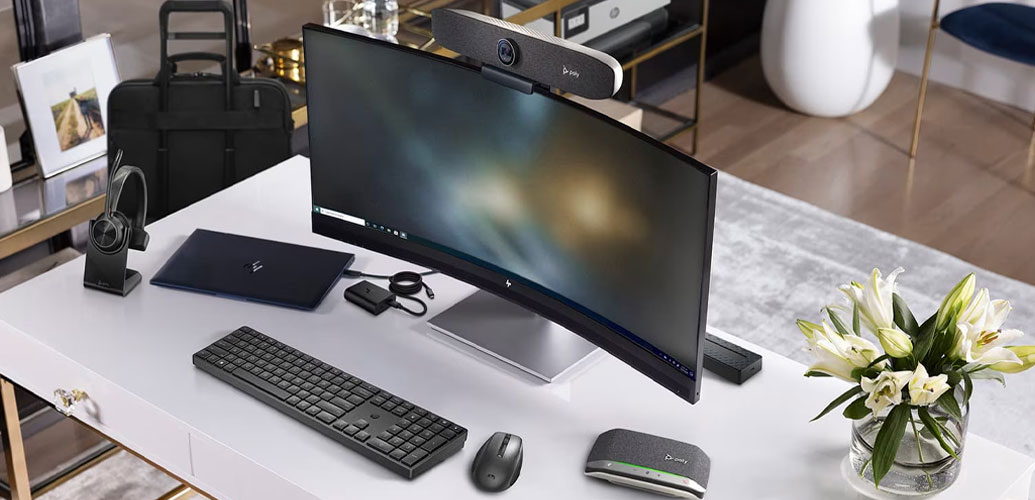 Curved monitor setup with laptop, keyboard, mouse, headphones, webcam, and vase of lilies on a white desk.