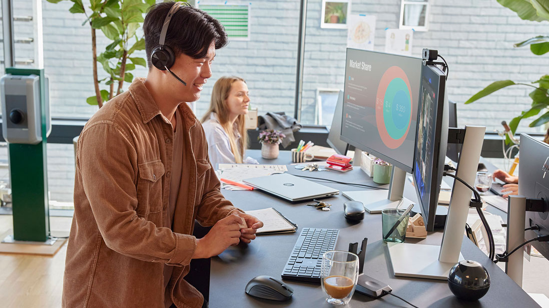 A man wearing headphones sits at a desk, focused on two computer monitors displaying various applications while an HP lapttop is kept on the desk.